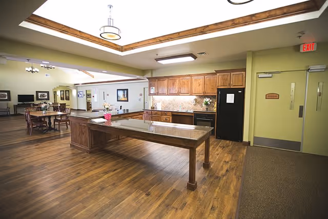 Interior view of a senior living facility kitchen and dining area with wooden floors, a large central island with a granite countertop, wooden cabinets, a black refrigerator, and a dining table with chairs in the background. The ceiling has recessed lighting and wooden beams, and there is an exit door on the right side.