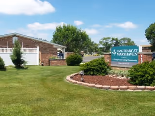 Outdoor view of the Sanctuary at Maryhaven senior living community sign surrounded by a landscaped garden with grass, bushes, and a tree. A brick building and white fence are visible in the background under a partly cloudy sky.
