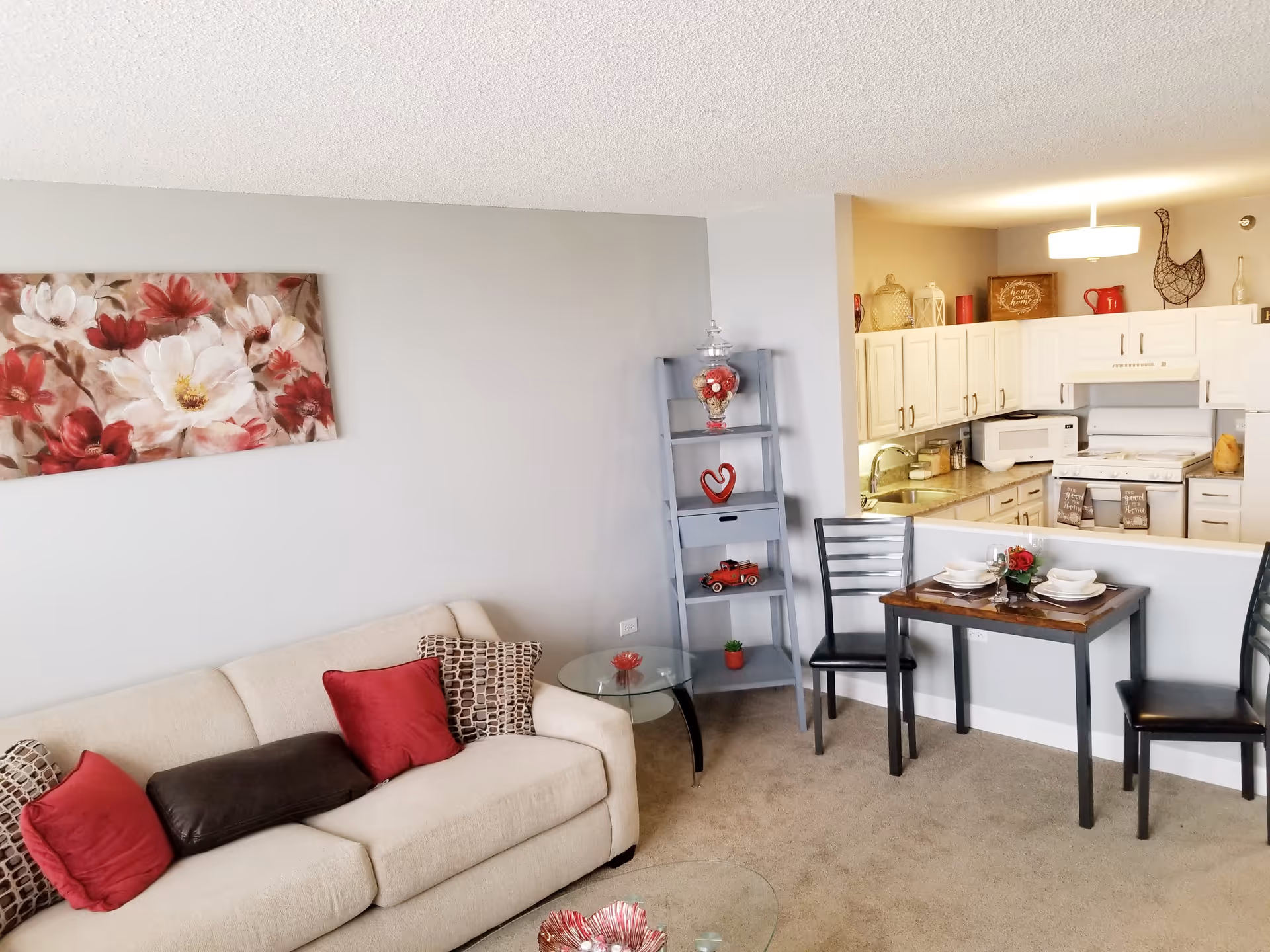 A cozy living area with a beige sofa adorned with red and patterned cushions, a glass side table, and a gray shelving unit decorated with various items. Adjacent to the living area is a small dining table set for two with black chairs. The background shows a white kitchen with cabinets, a stove, microwave, and decorative items on the countertop and shelves.