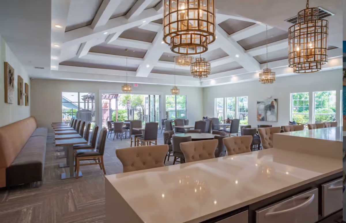 Spacious dining room with multiple tables and chairs arranged neatly. The room features a high ceiling with decorative beams and several large, cage-style pendant lights. Large windows and glass doors allow natural light to fill the space, and there is a long cushioned bench along one wall with individual tables and chairs opposite it. The foreground shows a polished countertop with upholstered chairs.