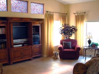 A cozy living room with a large wooden entertainment center holding a TV and stereo equipment, a red armchair with a cushion, a side table with a lamp and plant, and windows with yellow curtains letting in natural light.