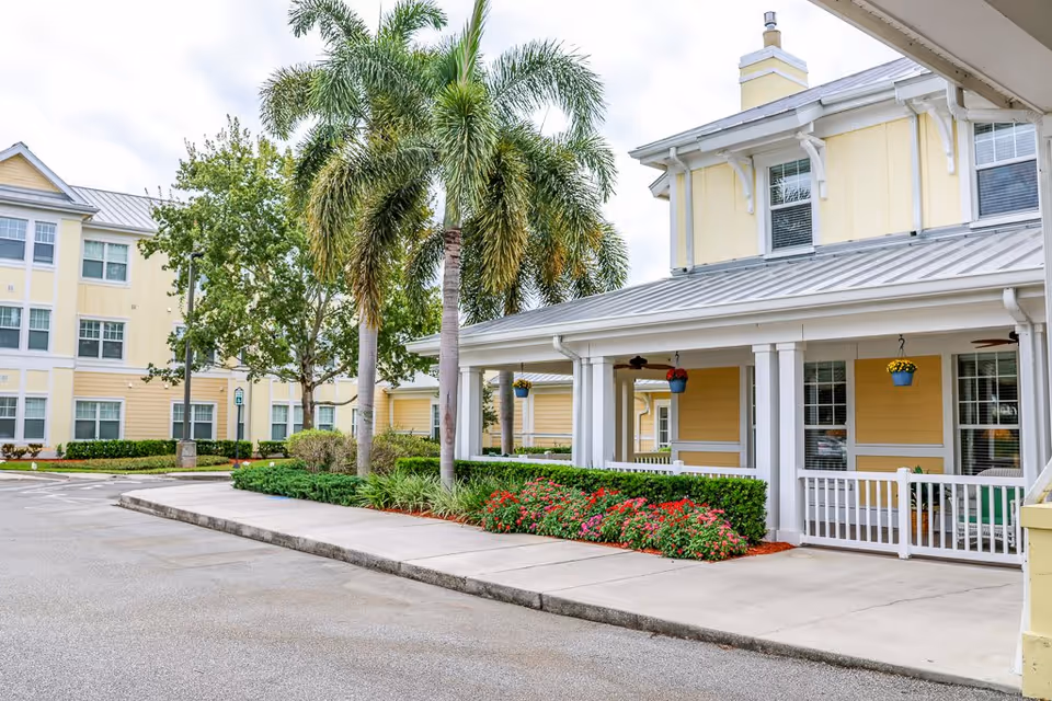 Exterior view of a senior living facility with yellow buildings, palm trees, and well-maintained landscaping including bushes and flowers along a sidewalk.