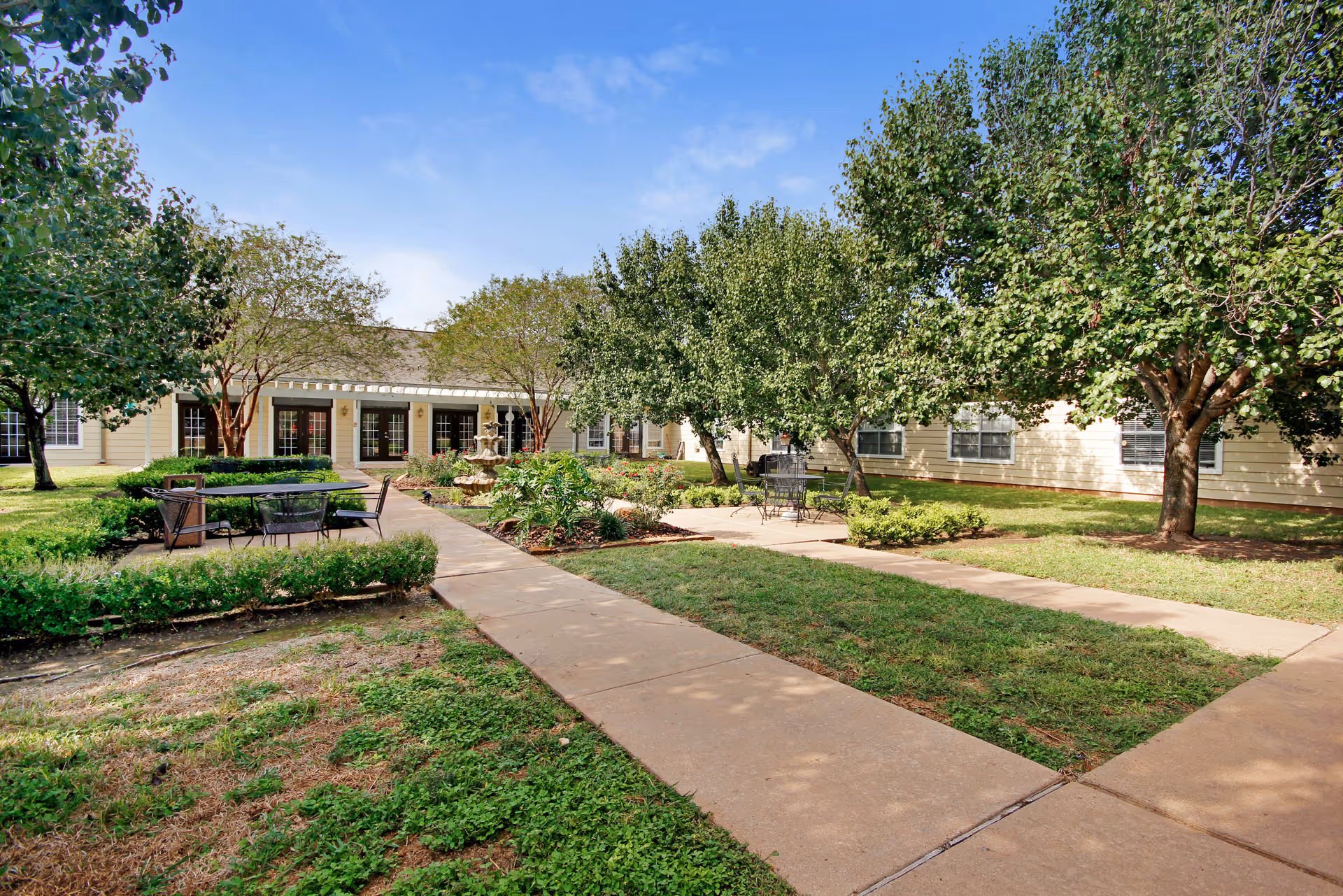 Courtyard with paved walkways, outdoor seating, trees, landscaping, and a single-story building facade in the background.