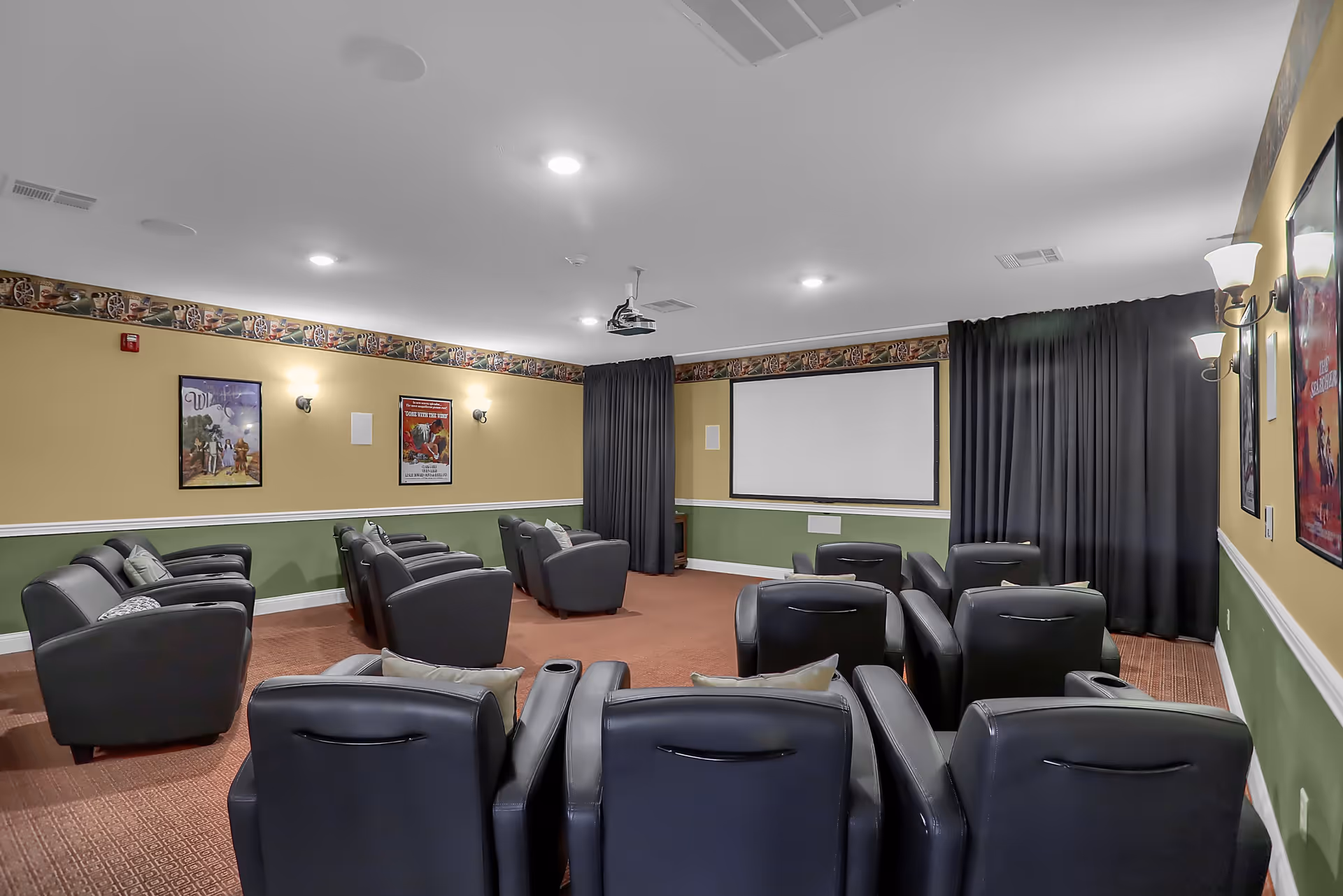 A small theater room with multiple rows of black leather recliner chairs facing a large white projection screen. The walls are painted yellow and green with decorative border trim near the ceiling, and movie posters are hung on the walls. The room is softly lit with wall sconces and ceiling lights, and dark curtains cover the windows.