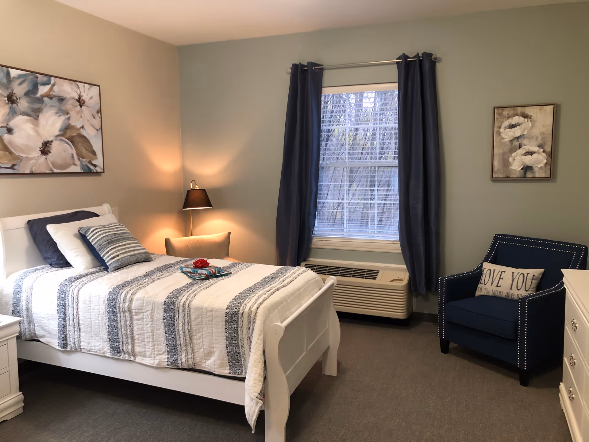 A cozy bedroom with a white bed frame and a striped blue and white quilt. The bed has multiple pillows, including a striped one and a dark blue one. There is a nightstand with a lamp emitting warm light next to the bed. A window with blue curtains and blinds is centered on the wall, with an air conditioning unit below it. To the right, there is a dark blue armchair with a pillow that says 'LOVE YOU'. The walls are painted in two tones, beige and light blue, and there are two floral paintings hanging on the walls.