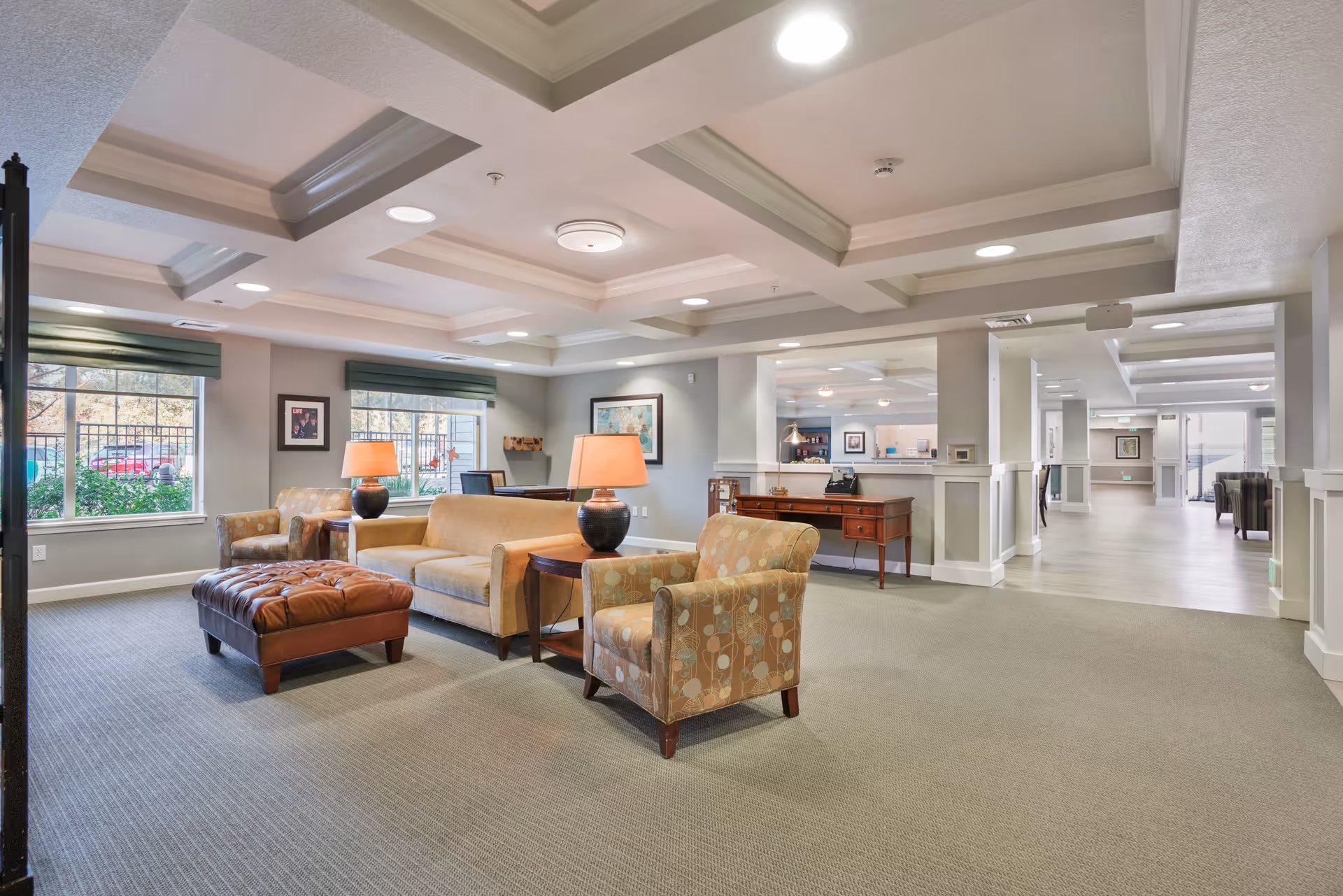 A spacious and well-lit living room area in a senior living facility with beige and patterned armchairs, a beige sofa, a brown leather ottoman, two table lamps with orange shades, and large windows with green valances. The room has a coffered ceiling with recessed lighting and a view into a hallway and reception area in the background.