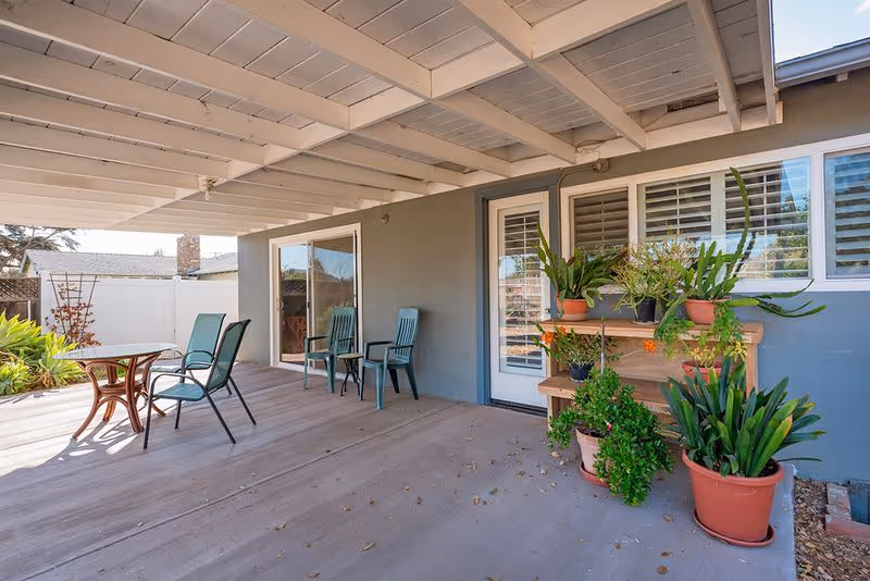 Covered patio with a table and chairs, potted plants on a shelving unit, and a sliding glass door into the house.