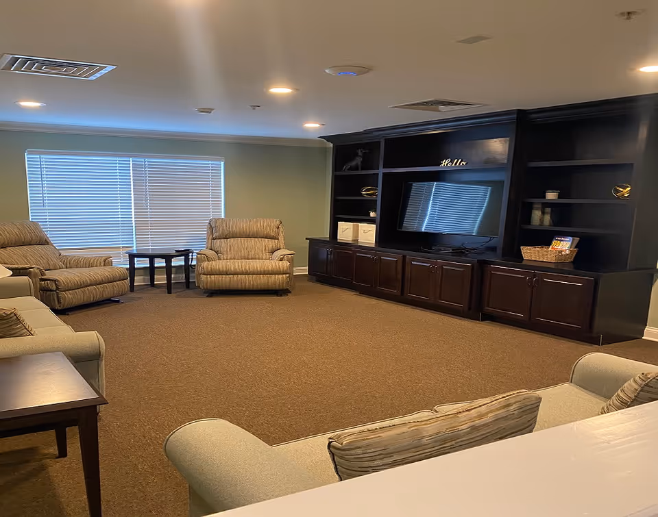 A cozy living room with beige recliner chairs and sofas arranged around a large dark wooden entertainment center with a flat-screen TV. The room has a large window with closed blinds and recessed ceiling lights.