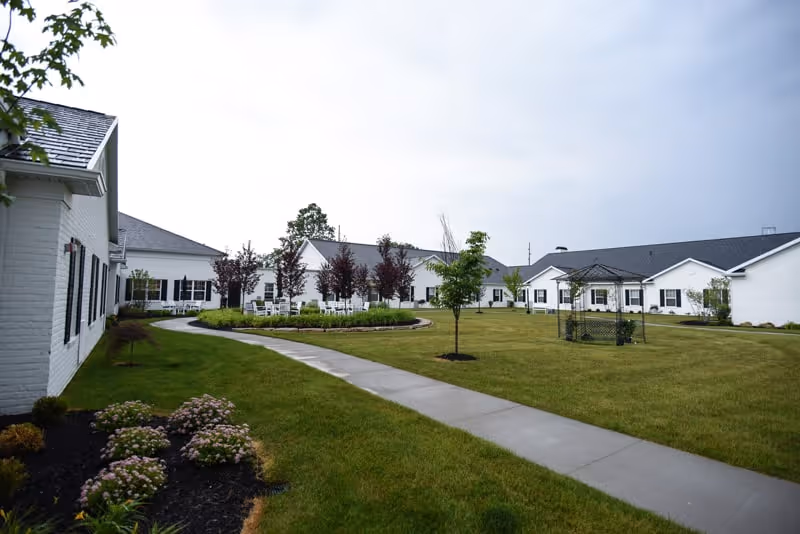 A spacious outdoor courtyard area of a senior living facility with a concrete walkway, green grass, small trees, flower beds, white buildings with black shutters surrounding the courtyard, and a small gazebo structure.