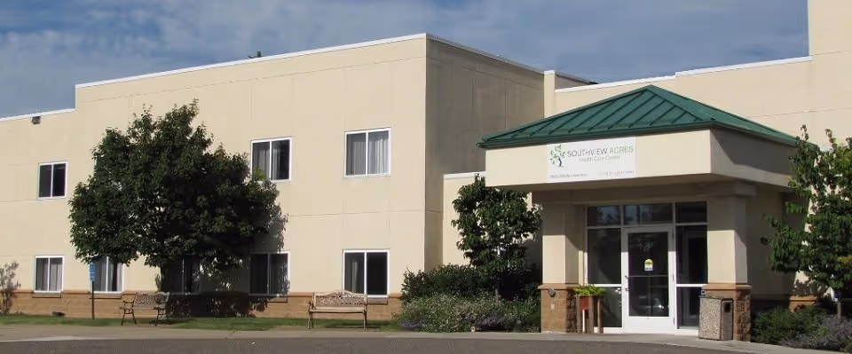 Front exterior of Southview Acres Health Care Center showing the building facade, main entrance with a green awning, windows, benches, and landscaping.