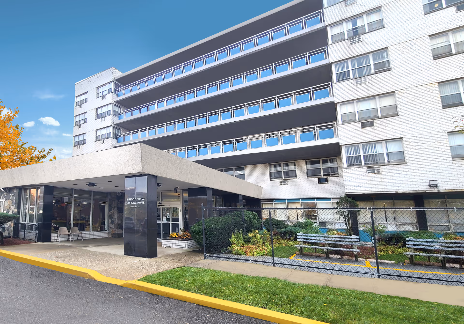 Entrance and facade of a multi-story white brick nursing home with a covered drop-off area and fenced courtyard.