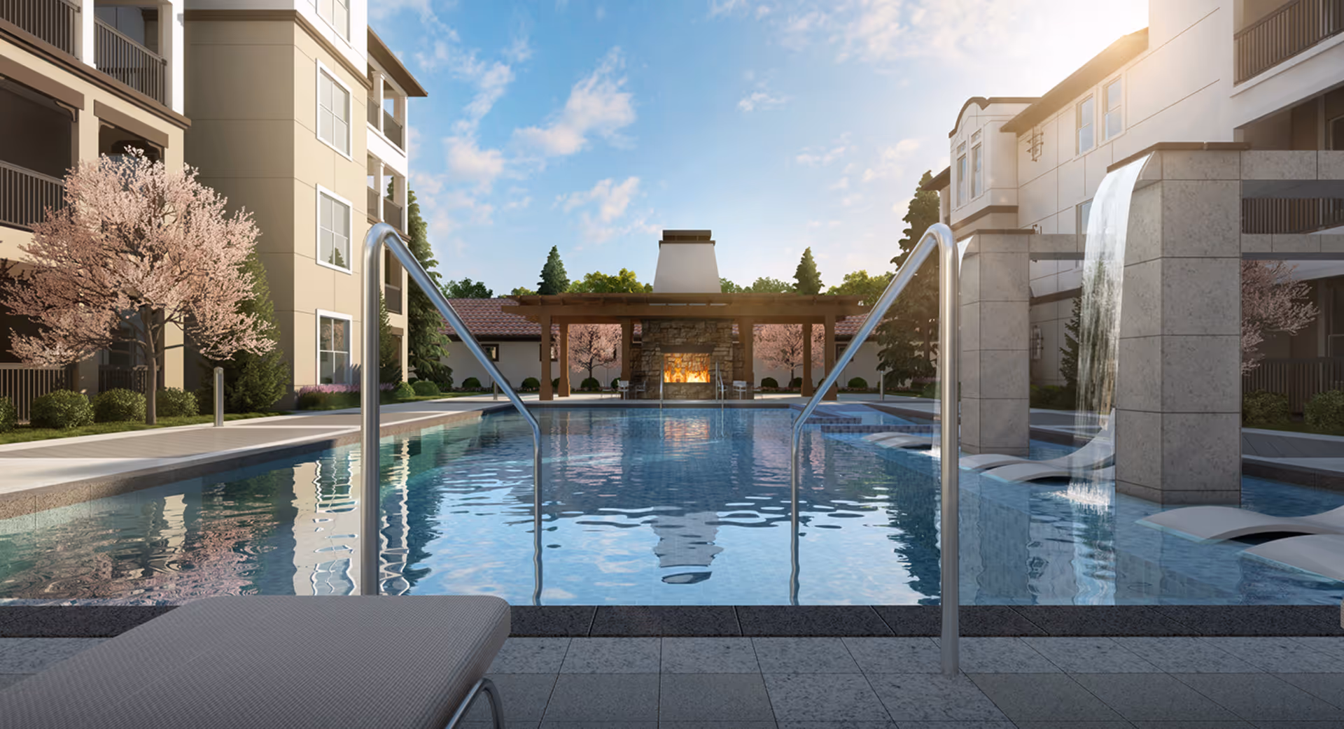 Outdoor swimming pool area at a senior living facility with metal handrails, a water feature cascading from a stone structure, lounge chairs, blooming trees, and a covered seating area with a fireplace in the background under a partly cloudy sky.