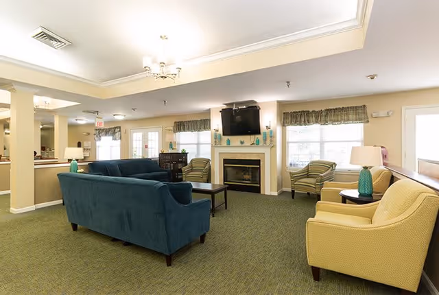 A spacious living room area with a green carpet, a blue sofa, two yellow armchairs, and two striped armchairs. There is a fireplace with a mounted TV above it, and windows with valances letting in natural light. The room is softly lit with ceiling lights and table lamps.