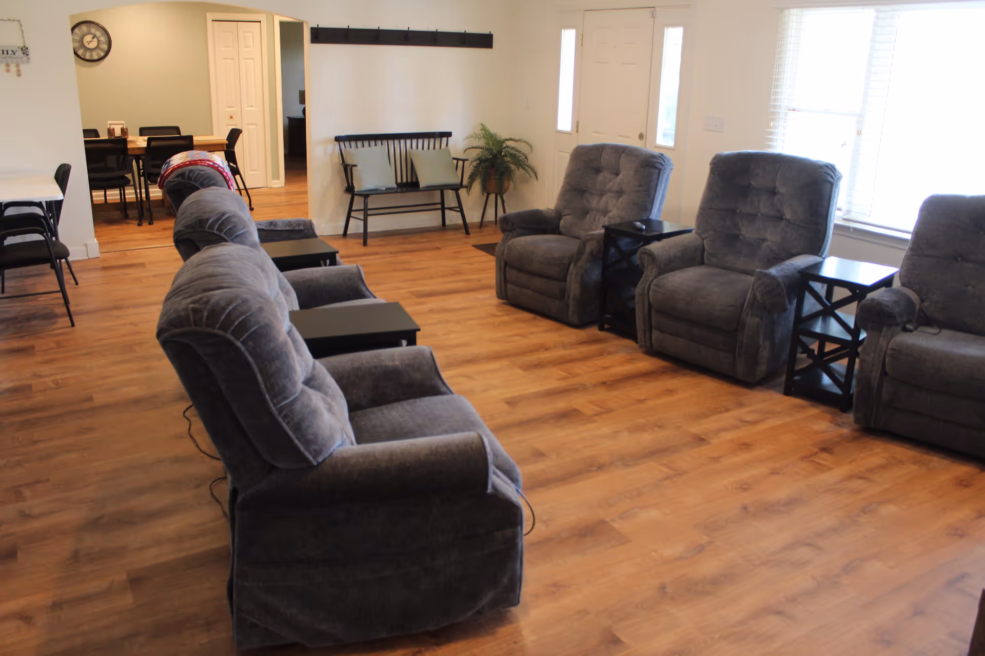 A bright common living room with several gray recliner chairs and side tables arranged on wood floors near a dining area.