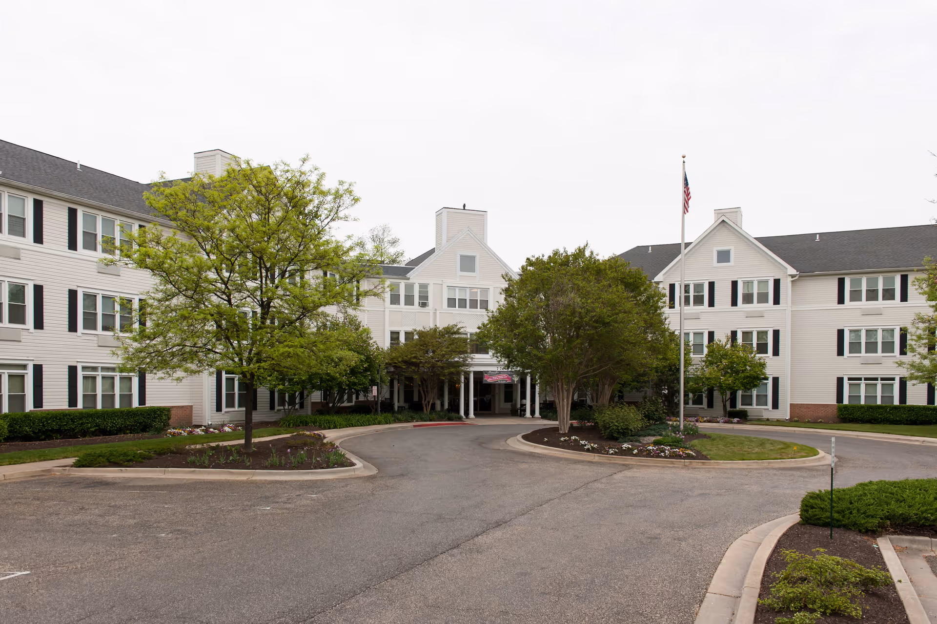Exterior view of Morningstar Assisted Living facility showing a large, three-story building with white siding and black shutters. There is a circular driveway with landscaped islands featuring trees and shrubs, and an American flag on a flagpole near the entrance.