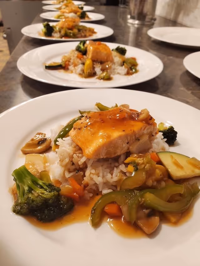 Close-up view of multiple plates lined up on a counter, each plate containing a serving of cooked salmon on a bed of white rice with mixed sautéed vegetables including broccoli, mushrooms, zucchini, and carrots, topped with a light sauce.