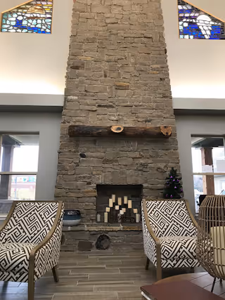 Interior view of a senior living facility lounge area featuring a tall stone fireplace with a wooden mantel. Two patterned armchairs are positioned in front of the fireplace, and a small decorated Christmas tree is placed on the right side of the hearth. Stained glass windows are visible above the fireplace, and large windows on either side allow natural light into the room.