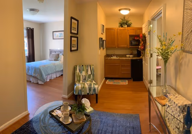 Furnished studio-style apartment showing a living area with a coffee table and chair, a small kitchenette, and a bedroom visible through an open doorway.