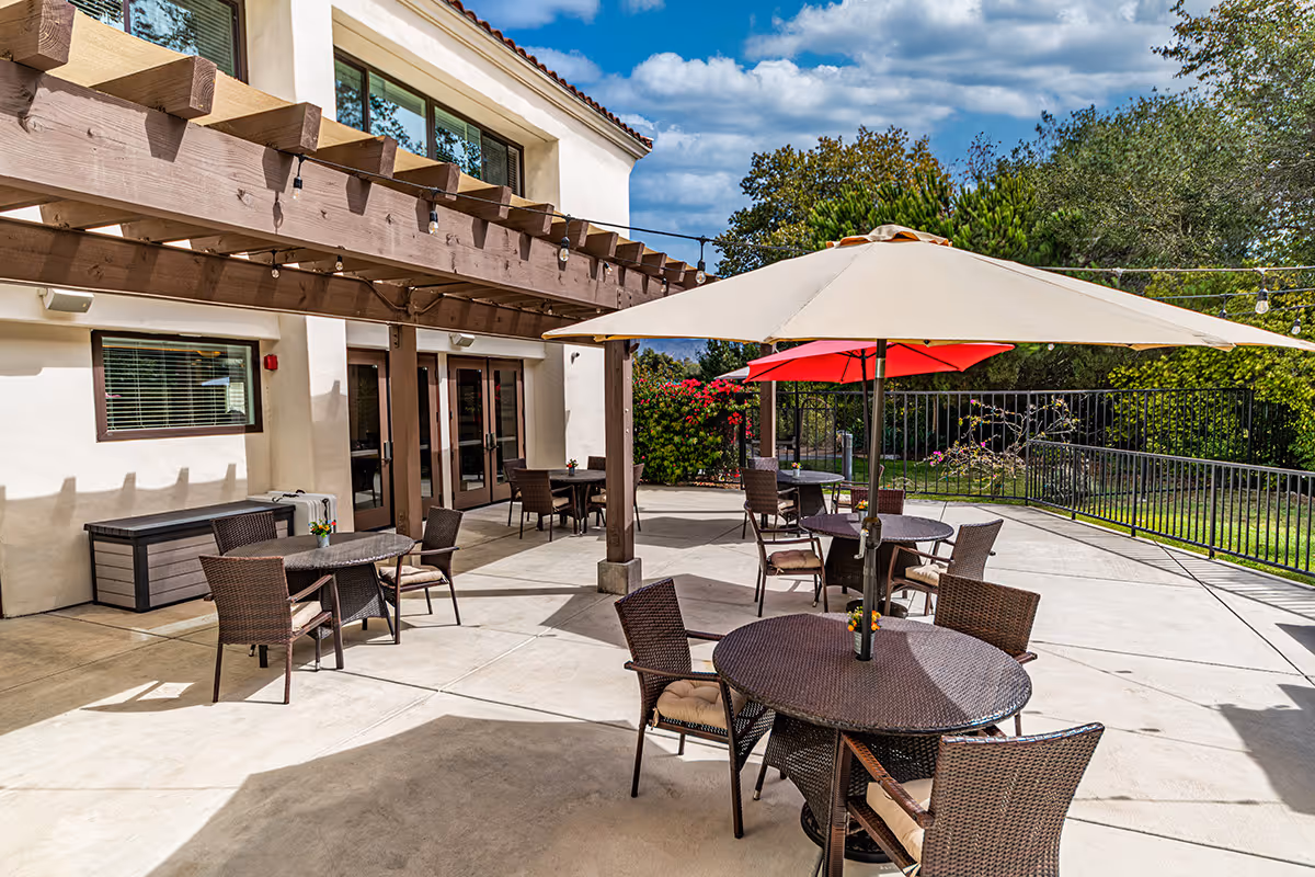 Outdoor patio area at a senior living facility with round wicker tables and chairs, some tables shaded by large umbrellas. The patio is adjacent to a building with large windows and a wooden pergola overhead. There are string lights hanging across the patio and greenery including trees and bushes surrounding the area.