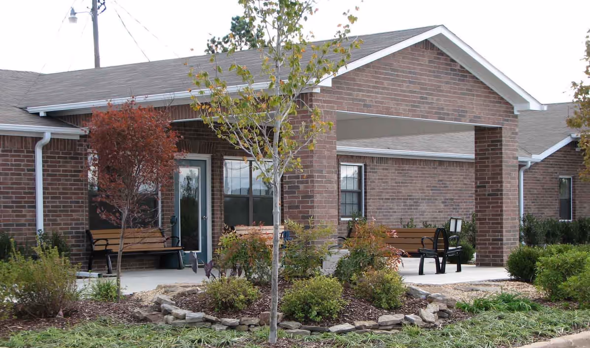 Exterior view of a brick building with a covered entrance supported by two brick pillars. There are benches on the porch area and small trees and shrubs planted in landscaped beds in front of the building.