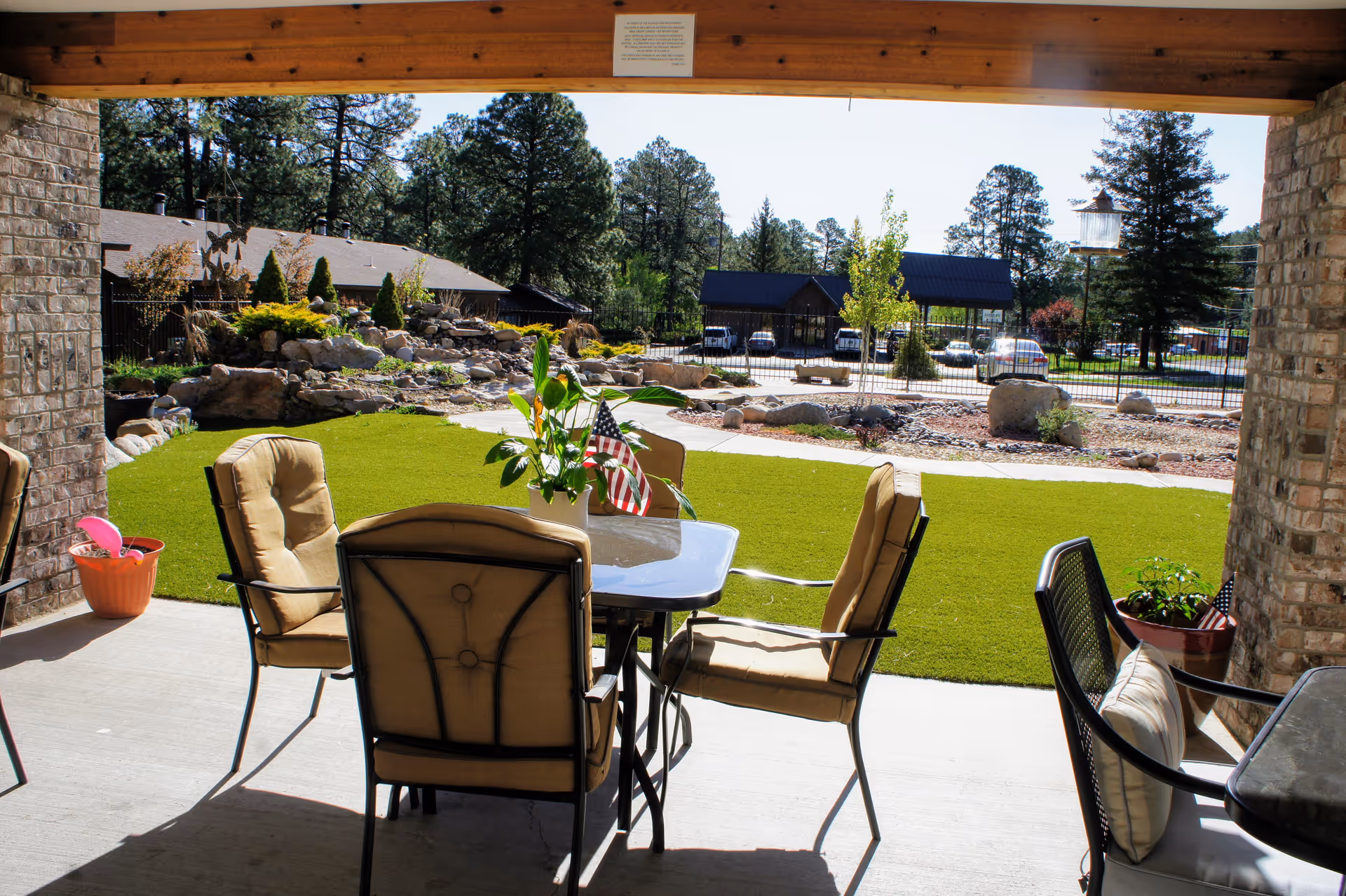 Outdoor patio area with a glass table surrounded by cushioned chairs. The patio overlooks a well-maintained lawn with landscaping rocks, plants, and trees. In the background, there is a parking area with several cars and a building. The scene is sunny with clear skies.