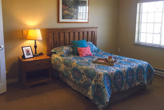 A cozy bedroom with a wooden bed frame and a colorful patterned bedspread. There are three decorative pillows on the bed, and a tray with a teapot and cup. A wooden nightstand with a lamp and a framed photo is next to the bed. A window with blinds allows natural light into the room.