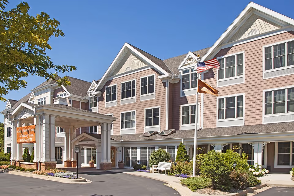 Front exterior of a multi-story senior living facility with a covered entrance, flags, and landscaped grounds.