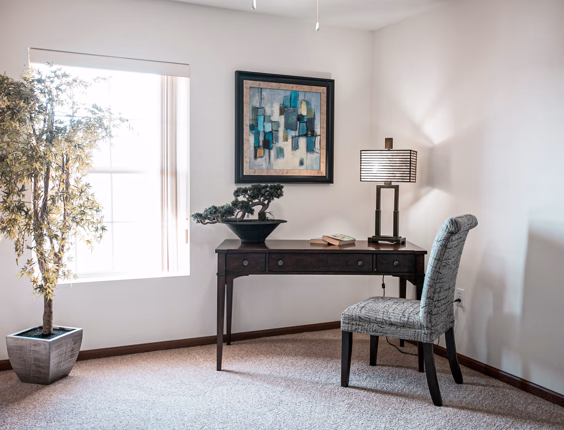 A small workspace in a room with a dark wooden desk, a patterned upholstered chair, a table lamp with a striped shade, a bonsai tree, two books, a large potted plant near a window with white blinds, and an abstract painting on the wall.