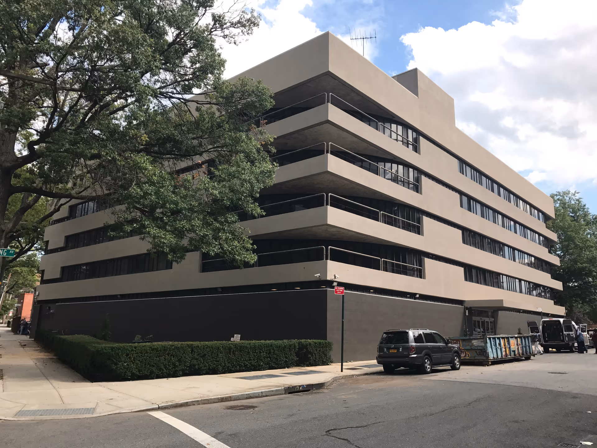 Exterior view of a multi-story beige and dark gray building with large windows and balconies, surrounded by trees and parked vehicles on the street corner.