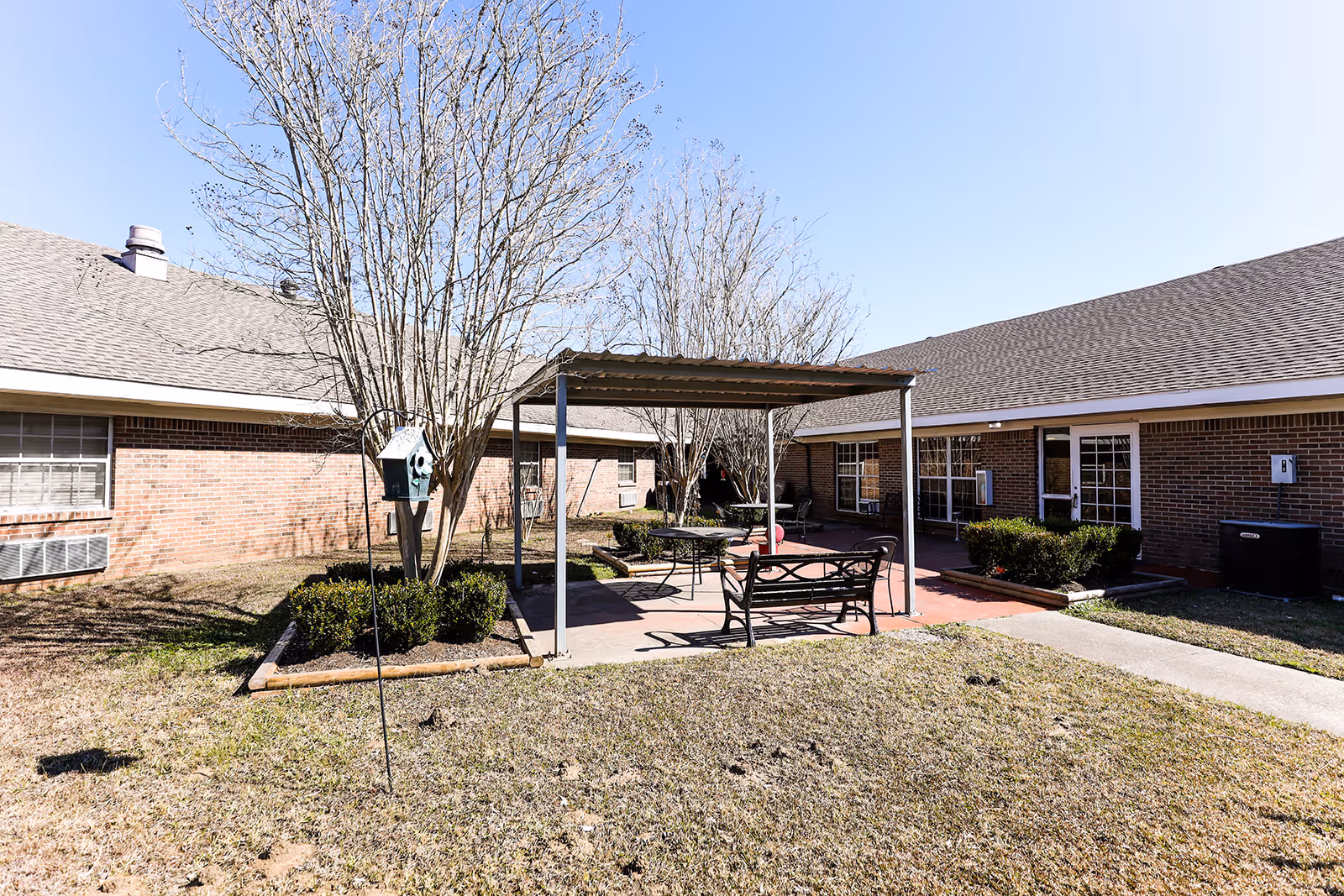 Outdoor courtyard area of a senior living facility with a covered seating area including benches and a table. The courtyard is surrounded by single-story brick buildings with windows and doors. Leafless trees and small bushes are planted around the seating area, and the sky is clear and blue.