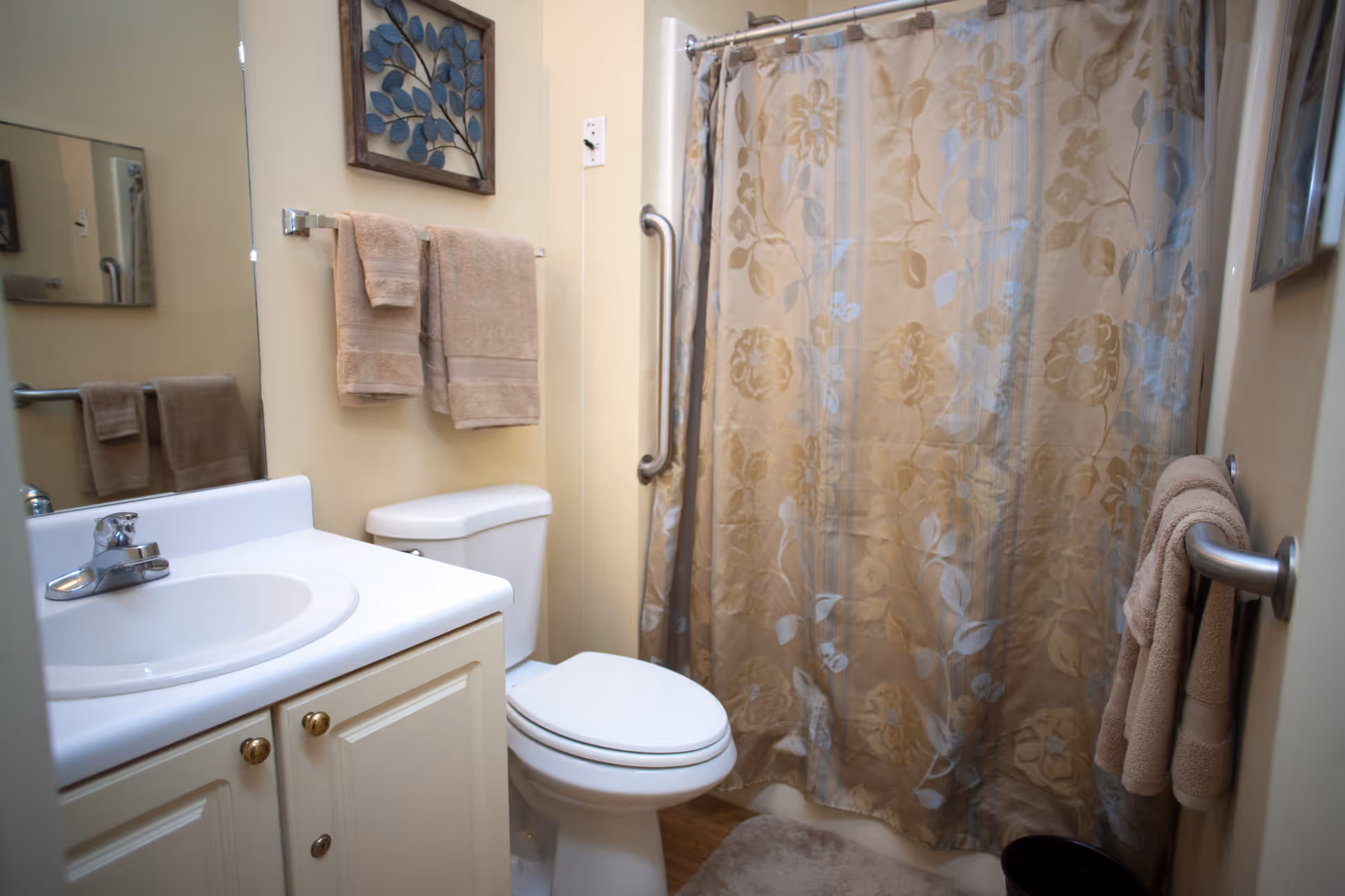 A small bathroom with a white sink and cabinet, a toilet, and a shower with a floral-patterned curtain. Beige towels hang on towel racks, and a framed artwork with blue leaves is mounted on the wall above the toilet.