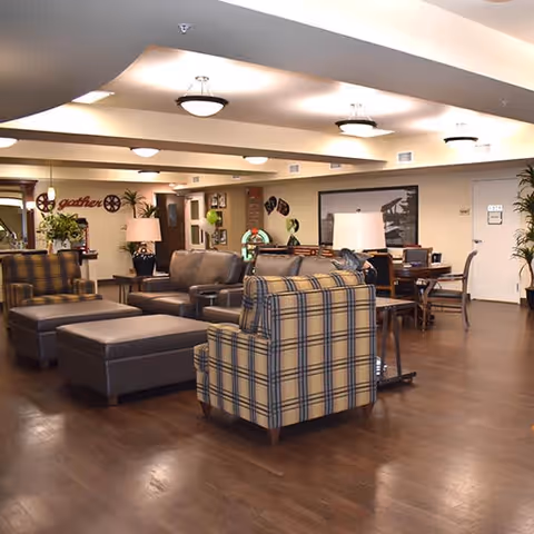 A spacious living room area with wooden flooring, featuring a plaid armchair, a leather sofa, a large ottoman, and several tables with lamps. The room is well-lit with ceiling lights and decorated with plants, wall art, and a sign that says 'gather'.