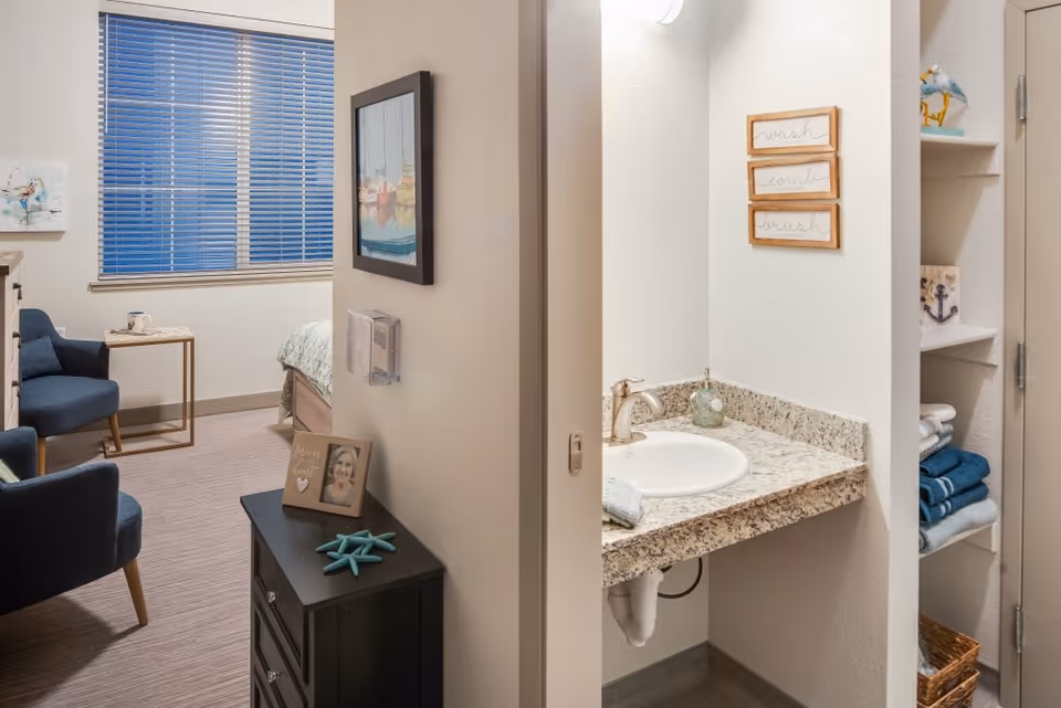 Interior view of a furnished bedroom with chairs and a bed visible through a doorway next to a small granite sink and open shelving.