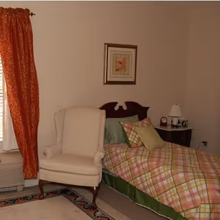 A bedroom with a single bed covered in a plaid bedspread with green, pink, and beige colors. There are multiple pillows on the bed. Next to the bed is a wooden nightstand with a white lamp, a clock, and some decorative items. A beige upholstered armchair is positioned near the bed. The room has beige walls, a framed picture above the bed, and a window with red patterned curtains. A carpet with a floral border is partially visible on the floor.