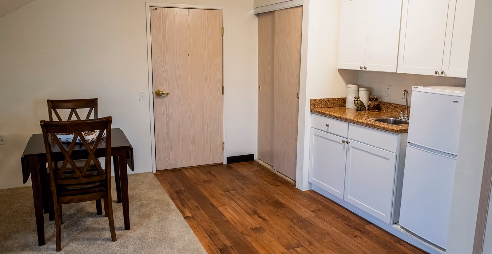Entryway of an apartment showing a small kitchenette with white cabinets and mini-fridge, a wooden dining table with two chairs, and the front door.