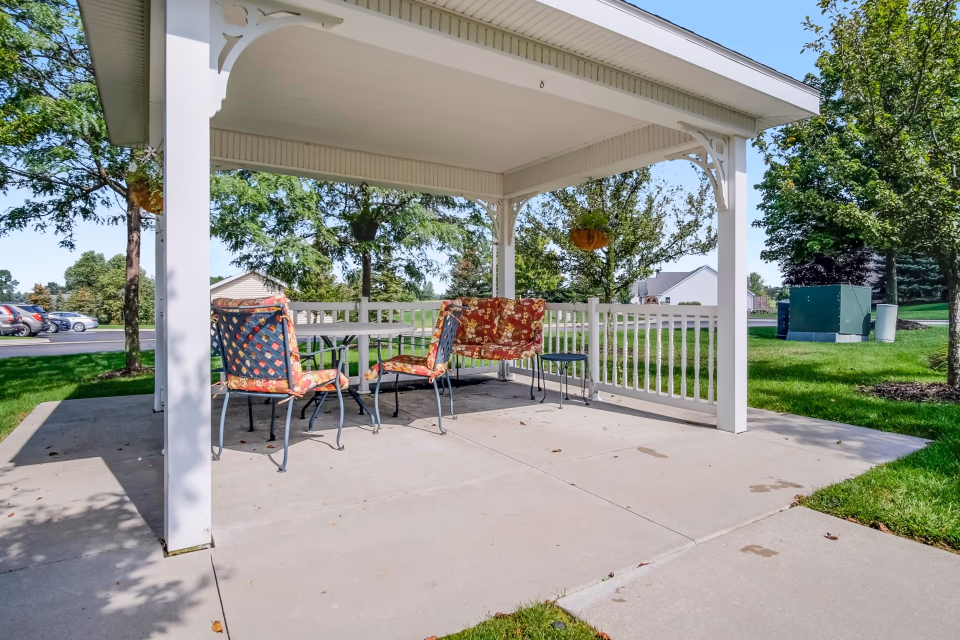 Outdoor covered seating area with a white pergola structure, featuring a table and four chairs with floral cushions, surrounded by green grass, trees, and a parking lot in the background.