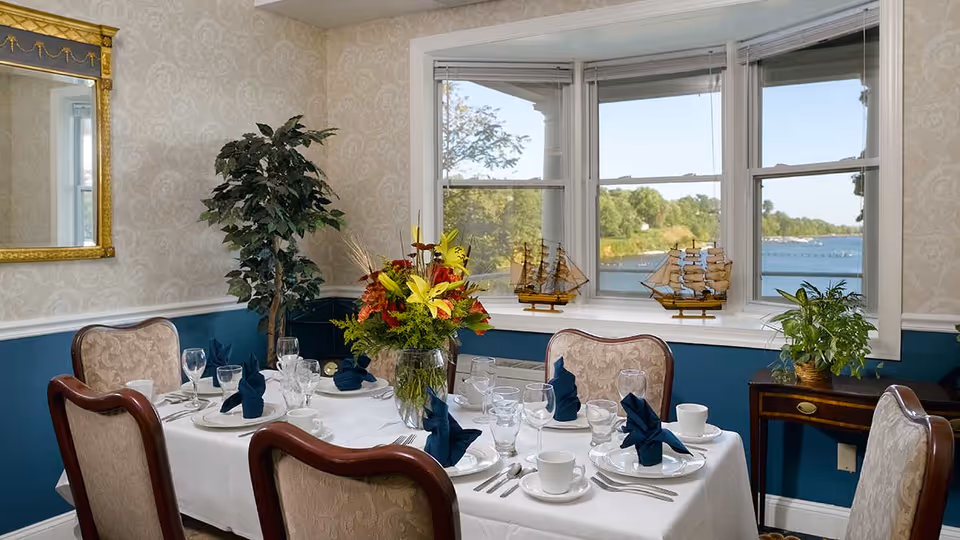 A formal dining room with a table set for six people, featuring white tablecloth, folded navy blue napkins, glassware, and white plates. A vase with colorful flowers is in the center of the table. The room has patterned wallpaper, a large window with a view of a lake and greenery outside, and decorative model ships on the windowsill. There is a potted plant in the corner and a small wooden side table with another plant.