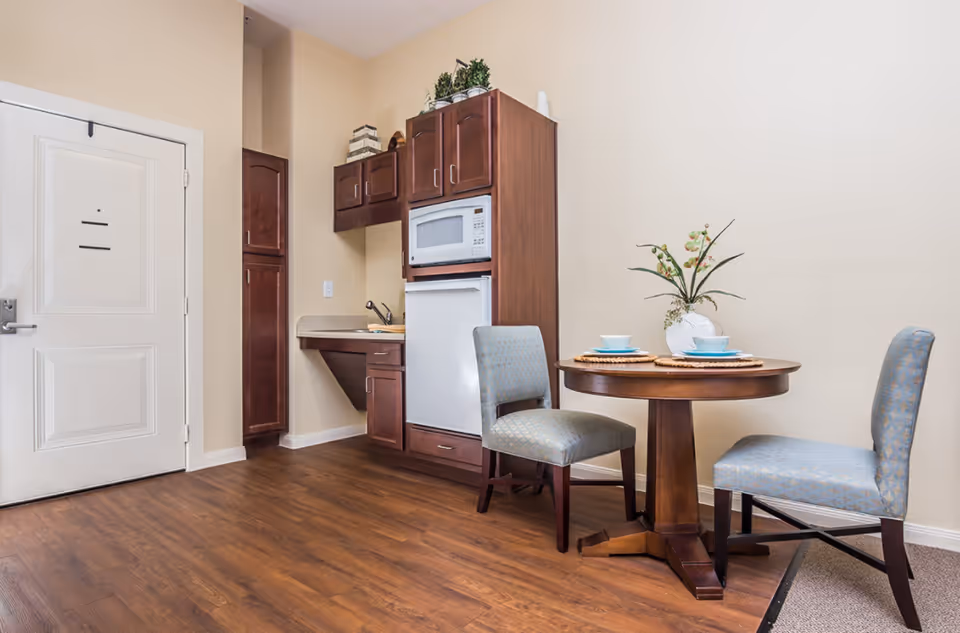 Small dining area with a round wooden table set for two with blue dishes and a white vase with flowers. Next to the table are two upholstered chairs with a blue patterned fabric. In the background, there is a kitchenette with wooden cabinets, a microwave, a small refrigerator, and a sink. The floor is wood, and the walls are painted beige.