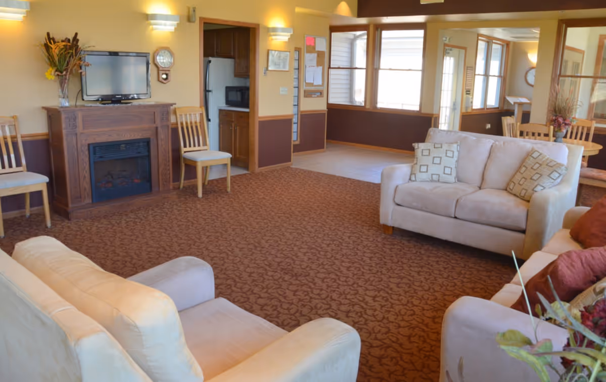 A bright communal living area with sofas and chairs, a TV over a fireplace, and a dining nook visible in the background.