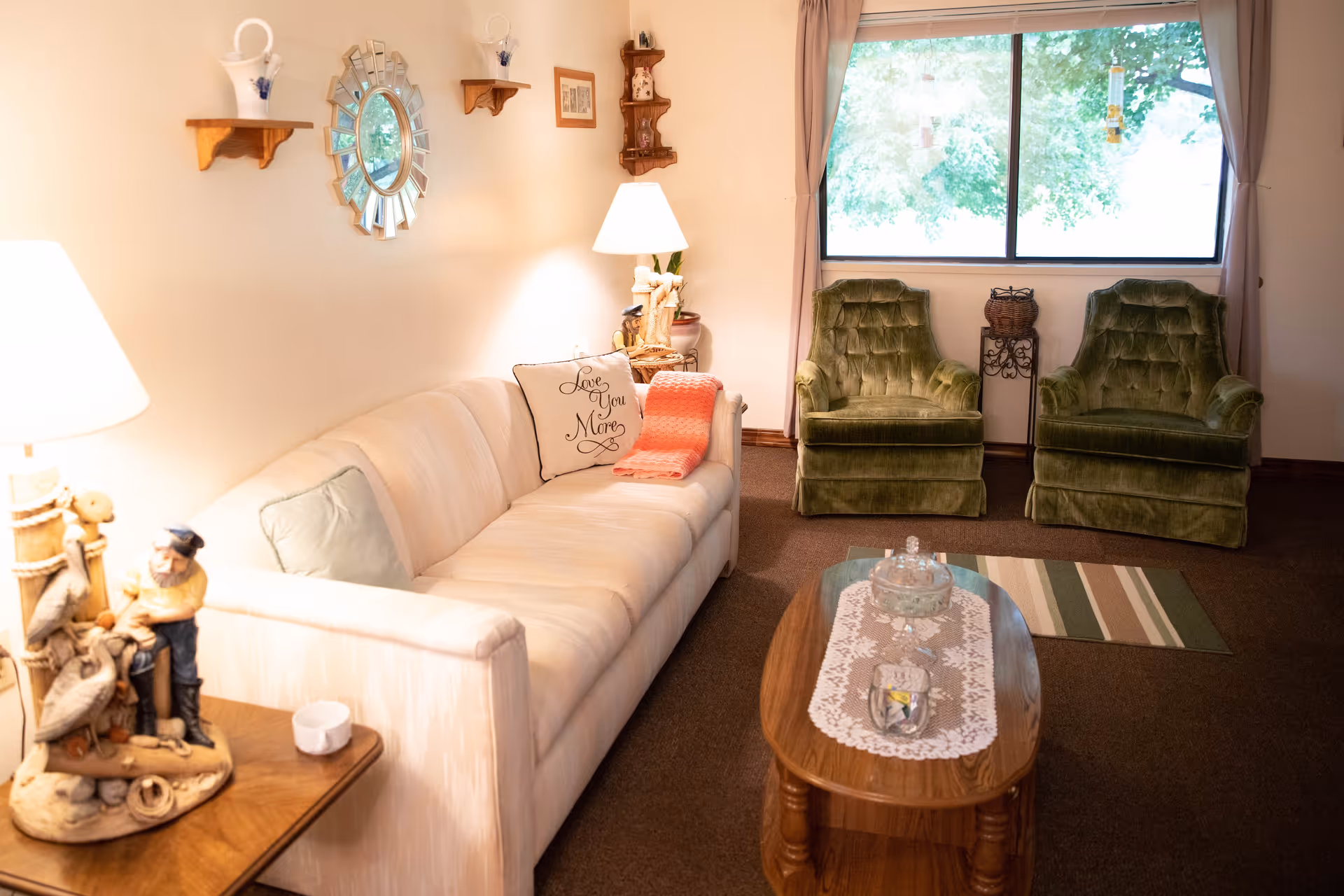 A cozy living room with a white sofa adorned with a pillow that says 'Love You More' and a coral throw blanket. Two green upholstered armchairs are placed near a window with beige curtains. A wooden coffee table with a lace runner and glass decorative items sits in front of the sofa. The walls have small wooden shelves with decorative items and a round mirror. A table lamp with a figurine base is on a side table next to the sofa.