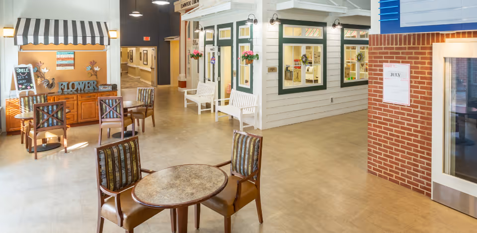 Interior view of a senior living facility common area with several small round tables and chairs arranged for seating. There is a decorative area with a striped awning, a wooden cabinet, and wall decorations including a sign that says 'SMILE It's a great day'. The hallway leads to other rooms, and there are white benches and flower pots hanging on the walls of a small house-like structure inside the facility. The floor is light-colored, and the walls are a mix of white paneling and red brick.
