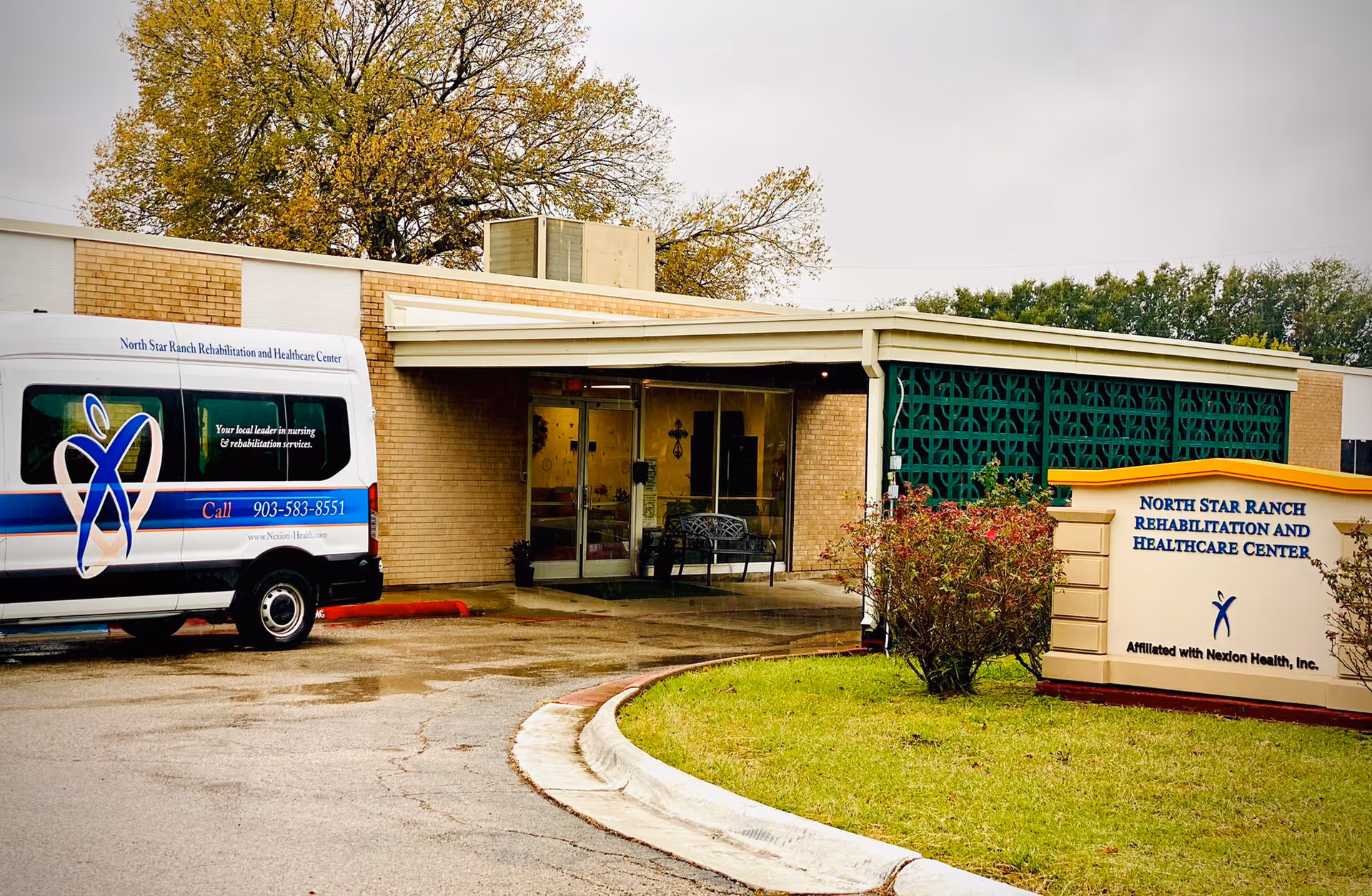 Exterior view of North Star Ranch Rehabilitation and Healthcare Center showing the entrance with a covered walkway, a parked facility van with branding, a sign with the facility name, and some greenery including bushes and a tree.