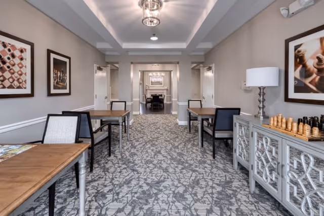 A well-lit interior room with patterned carpet and neutral-colored walls featuring framed chess-themed artwork. There are several wooden tables with chairs arranged along the sides of the room. On the right side, a decorative cabinet holds a chess set and a lamp. The room leads into another space with a dining table and chairs visible in the background.