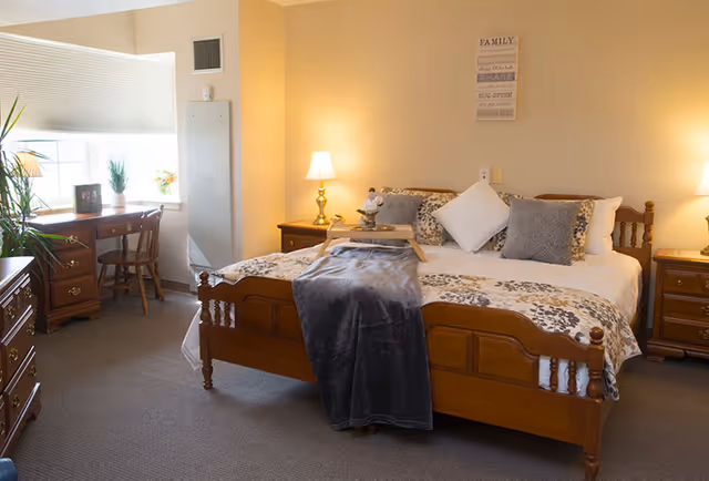 A cozy bedroom with a wooden bed frame, a neatly made bed with multiple pillows and a gray throw blanket. There are two wooden nightstands with lamps on each side of the bed. A wooden desk with a chair is positioned near a window with blinds, and there are plants and framed photos on the desk. The walls are painted a soft beige color, and a decorative sign hangs above the bed.