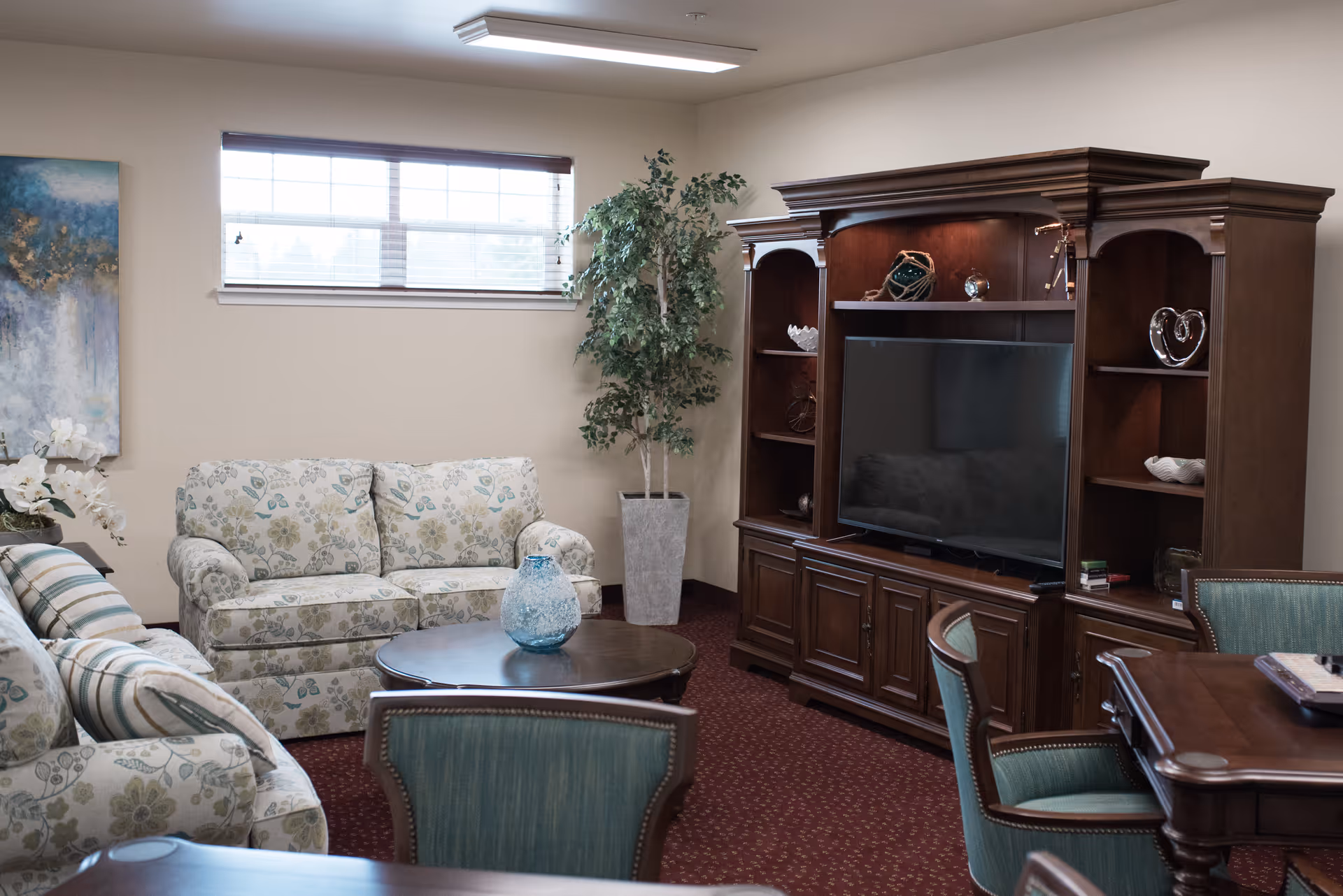 A cozy living room with floral patterned sofas, a round wooden coffee table with a blue decorative vase, a large wooden entertainment center with a flat-screen TV, and a tall potted plant near a window with blinds.