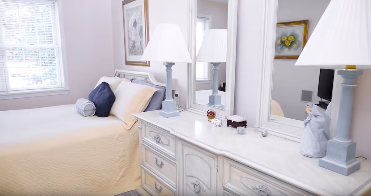 Sunlit bedroom with a neatly made bed next to a white dresser topped with twin lamps and a large mirror.
