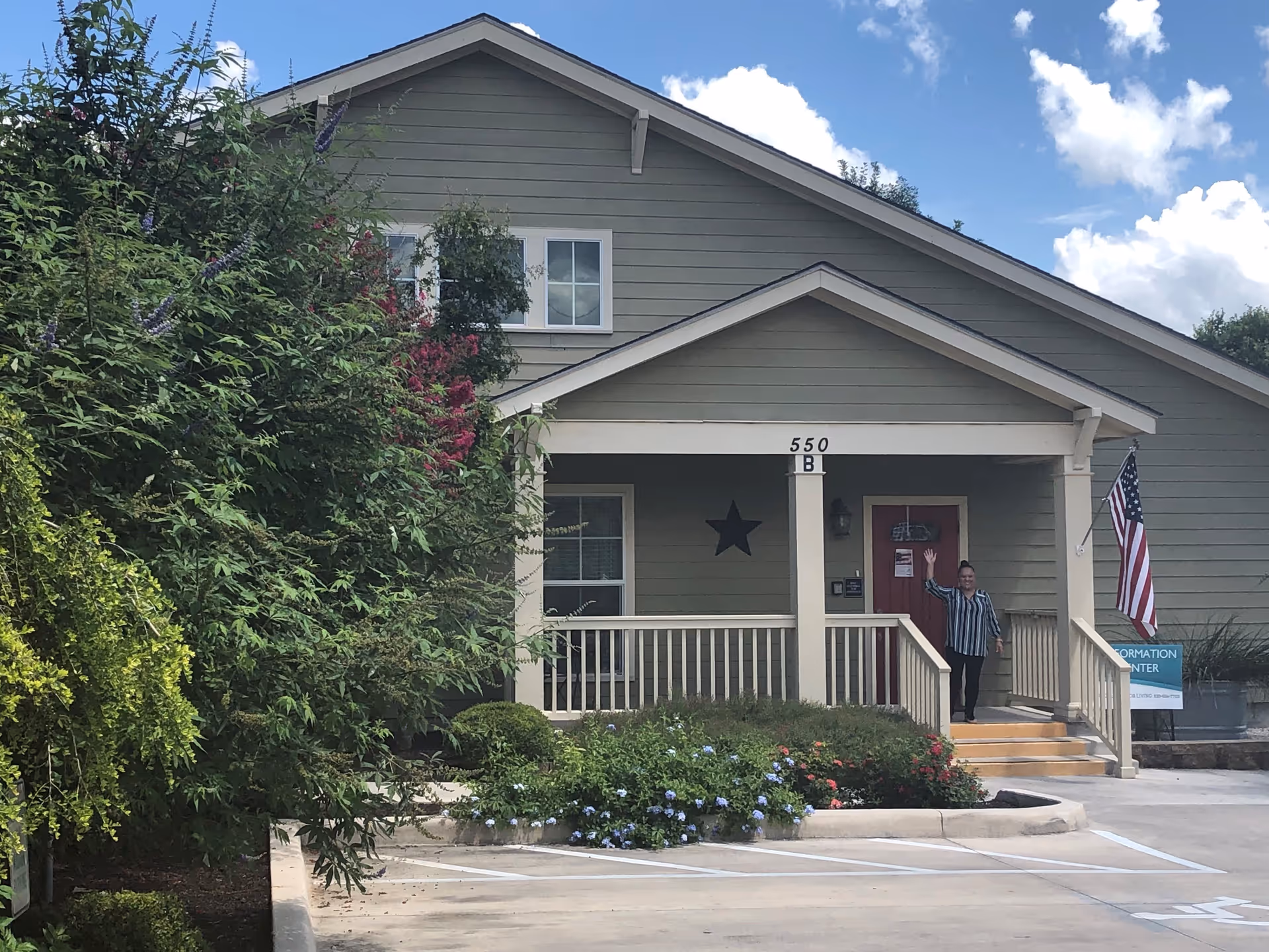 Exterior view of a single-story building with a porch, beige siding, and a red door. A person is standing on the porch waving. There is an American flag on the right side near the entrance and various bushes and flowers in front of the building. The sky is partly cloudy.