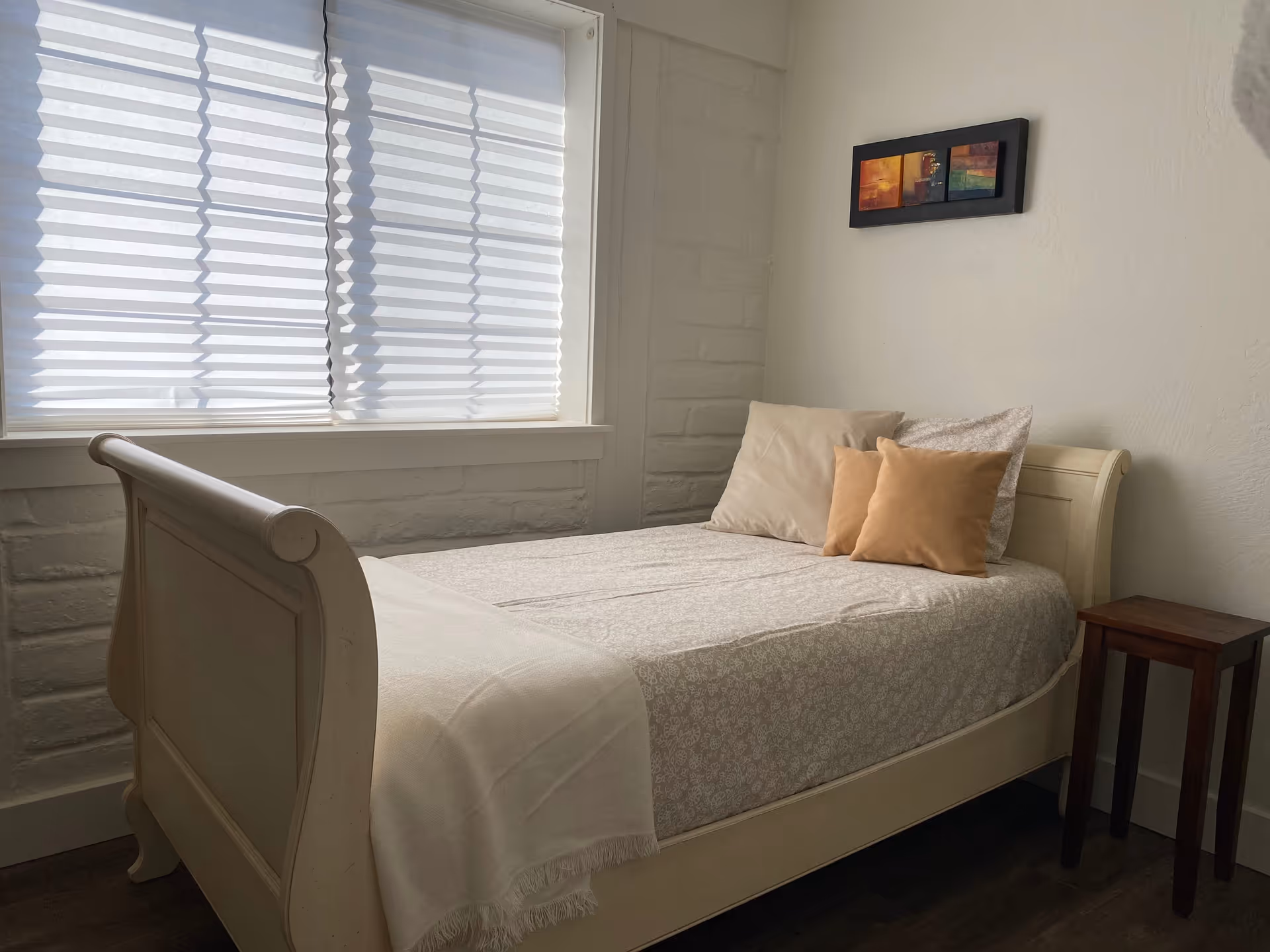 A small bedroom with a single bed against a white brick wall. The bed has light-colored bedding with three pillows in beige and light brown shades. There is a wooden side table next to the bed and a window with white blinds letting in natural light. A small framed abstract painting hangs on the wall above the bed.