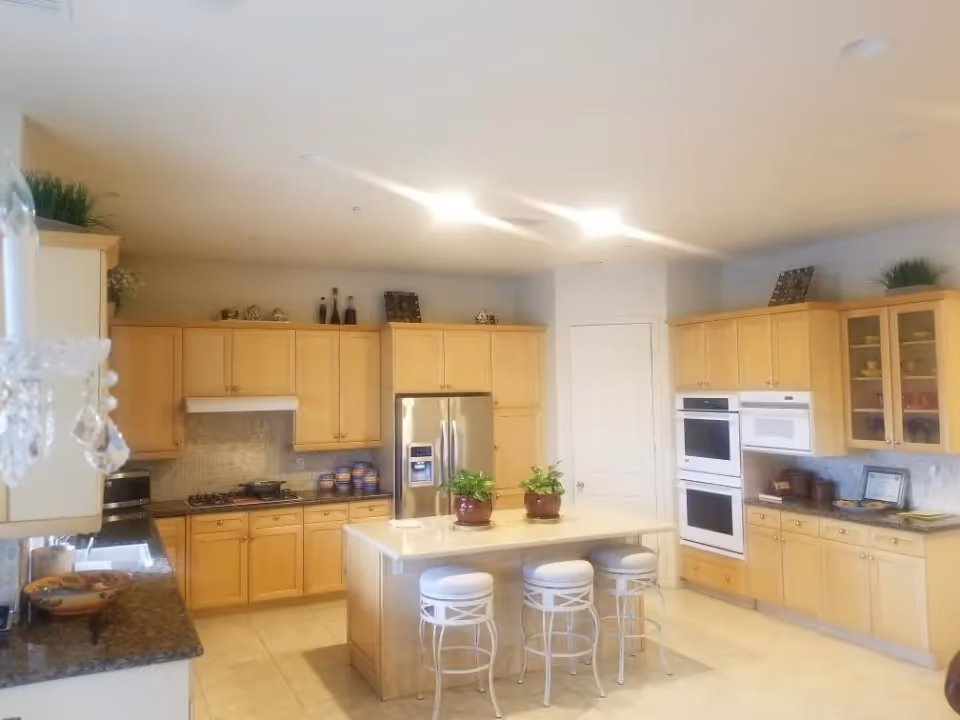 A spacious kitchen with light wood cabinets, a central island with three white stools, and stainless steel appliances including a refrigerator and double oven. The countertops are granite, and there are decorative plants and items on top of the cabinets and island. The floor is tiled, and the kitchen is well-lit with ceiling lights.