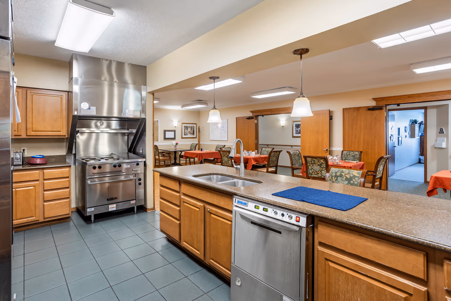 Interior view of a kitchen area in a senior living facility with wooden cabinets, a stainless steel stove and dishwasher, a double sink, and a countertop. Beyond the kitchen is a dining area with tables covered in red tablecloths and chairs with floral upholstery. The room is well-lit with ceiling lights and pendant lamps.